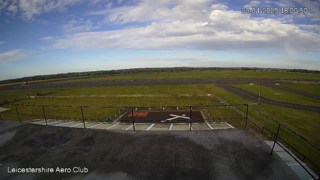 A wide view of a grassy runway at the Leicestershire Aero Club is captured. In the foreground, a helipad marked with a white cross sits on a dark surface. The area is bordered by a fence, and there are scattered markings on the grass. The sky above is partly cloudy with patches of blue, and the landscape extends into the distance, revealing a flat, open terrain with an additional runway visible. The time and date displayed on the image indicate it is late afternoon.