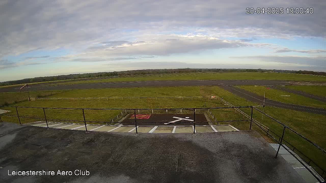 A view from a webcam at the Leicestershire Aero Club, showing a wide expanse of green grassland, a runway, and a few scattered markings. In the foreground, there are railings along the edge of a building. The sky is partly cloudy with patches of blue. A landing strip is visible, with a few signs and a cross marking an area on the ground. The scene conveys a serene environment typical of an airport facility.