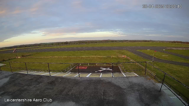 The image shows a panoramic view of a grassy airfield with a clear sky. In the foreground, there is a black and white "X" marking on the ground, indicating a landing area. Nearby, a fence runs along the edge of the field, and a vertical flagpole can be seen with a flag flying. The background features a long stretch of runway, and beyond that, green fields and a distant horizon. The date and time in the corner indicate the image was captured on April 20, 2025, at 20:00:40.