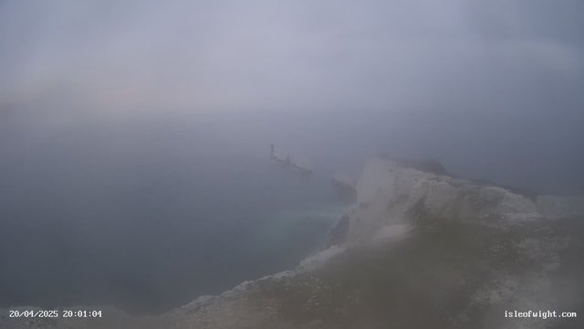A foggy coastal scene showing a rocky cliff leading down to the sea. In the background, a silhouette of a distant ship is partially visible through the mist. The sky appears overcast and muted, blending into the foggy water. The image conveys a sense of calmness and isolation in a cloudy environment.