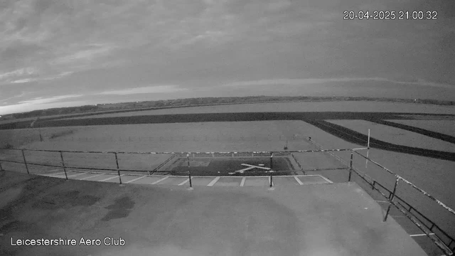 A black and white webcam image of a landscape under a cloudy sky. There is a large expanse of open land with distinct lines marking a runway and the edge of the aerodrome in the foreground. A low fence appears to surround the area. The image captures a quiet, still atmosphere, with no visible people or aircraft. The timestamp in the corner indicates the image was taken on April 20, 2025, at 21:00:32.