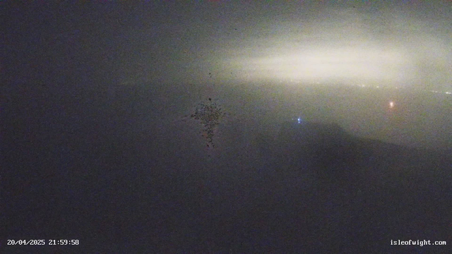 A foggy scene captured by a webcam, with a hazy view obscuring most details. The background features a soft, diffuse light suggesting distant lights or reflections. A faint cluster of shapes appears in the center, possibly representing objects or light sources. The overall atmosphere is dark and unclear, indicating limited visibility. The date and time are displayed in the lower left corner.