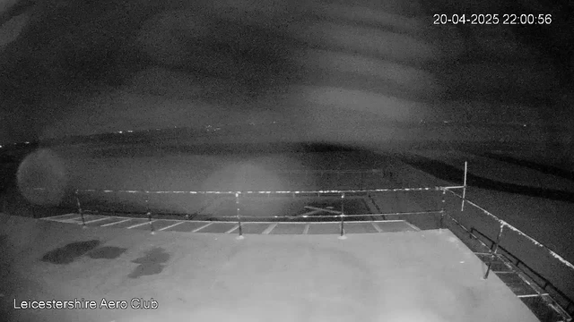 A nighttime view from a webcam at the Leicestershire Aero Club. The scene is mostly dark with some faint lights visible in the distance, suggesting the presence of a town or city beyond the airport. In the foreground, there is a flat surface with a railing along one edge, and some indistinct markings on the ground. The overall atmosphere is quiet and still, with minimal visible detail due to the low light conditions. There are soft, blurred patches across the image, indicating possible fog or moisture on the lens. The date and time at the top of the image read April 20, 2025, at 10:00:56 PM.