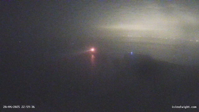 A dimly lit scene features a body of water with a faint reflection. In the center, there is a small, bright orange light, possibly a buoy or a distant boat. To the right, there is a smaller blue light. The background is mostly obscured by fog or low visibility, creating an atmospheric and somewhat mysterious setting. The timestamp at the bottom indicates the date and time of the image.
