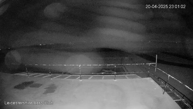 A night-time view from a webcam at Leicestershire Aero Club. The image is mostly dark, with a blurred foreground showing a railing and a portion of a flat surface. In the background, there are faint lights visible in the distance, suggesting urban area or reflections possibly from other objects. The overall image has a grainy appearance. The timestamp in the corner indicates the date and time as April 20, 2025, at 11:01 PM.
