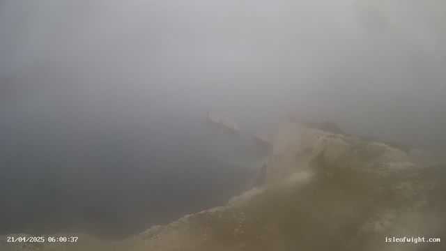 A foggy coastal scene with blurred outlines of rocky cliffs and distant structures partially obscured by mist. The water appears calm and dark, reflecting the hazy sky. The overall atmosphere is muted and gray, suggesting low visibility conditions. A timestamp at the bottom indicates the date and time of the image.