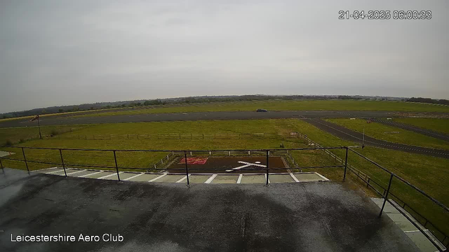 A view from a webcam at Leicestershire Aero Club, showing a large, open grassy area with a runway extending into the distance. The sky is mostly cloudy and gray. In the foreground, there is a wooden fence and a helipad marked with a white cross. To the right, a road intersects the grassy area. There is no visible activity on the runway. The image is timestamped at 6:00:33 AM on April 21, 2025.