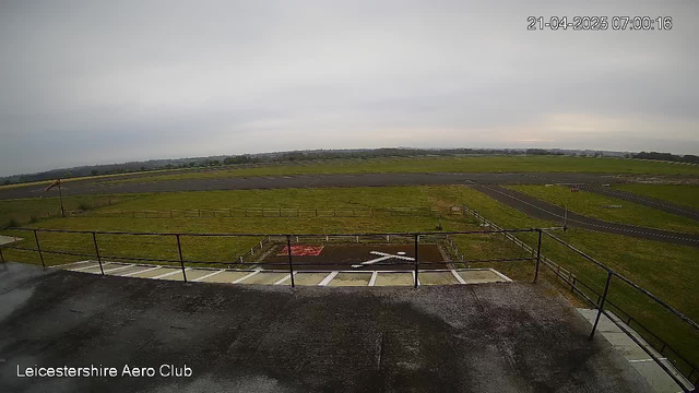 A view from a webcam at the Leicestershire Aero Club. The image shows a wide expanse of green grass and a long runway extending into the distance. A few white markings are visible on the ground, which likely indicate runway markings. The sky is overcast with gray clouds, suggesting a cloudy day. In the foreground, there is a railing and part of a building structure, which appears to be a rooftop view.
