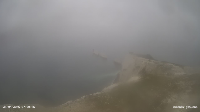 A foggy scene near a coastline. The view is obscured by dense mist, making it hard to see details. In the foreground, there is a rocky shoreline, with the edge of the land visible. In the background, faint silhouettes of what appear to be structures or boats are discernible but are mostly shrouded in fog. The overall atmosphere is muted and blurry, with a feeling of calmness.