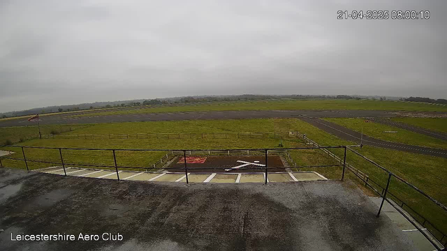 A view from a high vantage point overlooking a grassy airfield on a cloudy day. In the foreground, there is a gray, flat rooftop with a railing. Below, a large, paved runway stretches horizontally, bordered by grass. A white cross shape is marked on the ground, along with a red area. In the distance, there are trees lining the horizon, and the sky appears overcast, creating a dull, muted atmosphere.