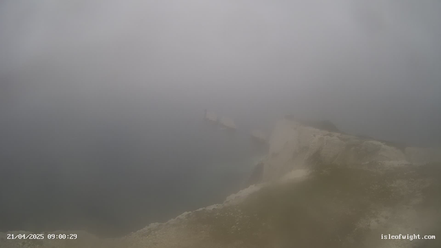 A foggy landscape is depicted, with an indistinct view of a coastline meeting a body of water. The scene is shrouded in mist, making visibility very low. Some rocky formations are slightly visible in the foreground, while the background merges into the gray fog. The overall atmosphere appears tranquil yet obscured. A timestamp at the bottom indicates the date and time of the image.