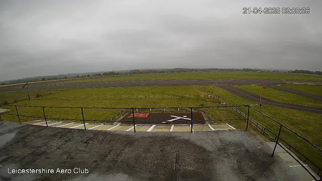 A cloudy sky overlooks a green field with an airport runway. In the foreground, there is a railing on a building with a viewing area. An 'X' marking is visible on the ground, indicating a designated area. The runway is partly visible, and the landscape extends into the distance with low hills. The scene gives a sense of an aerial view from the Leicestershire Aero Club.