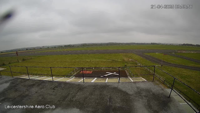 A view from a webcam positioned at the Leicestershire Aero Club showing a large, grassy field and an airstrip in the distance. The sky is overcast and gray, indicating cloudy weather. There is a marked area on the ground that features a large white X surrounded by a red square, and a section of the airstrip appears to be visible in the center of the image. The foreground shows a metal railing along a flat surface. The atmosphere is quiet and serene, typical of an aviation facility.