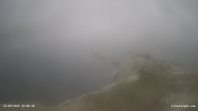 A foggy coastal scene featuring a rocky shoreline that gradually fades into a blurred ocean view. Several indistinct shapes of boats are visible in the water. The atmosphere is misty and gray, creating a sense of mystery and stillness. The date and time at the bottom left corner indicate April 21, 2025, at 10:00:18.