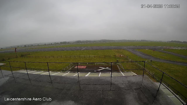 A cloudy and overcast view from a webcam at Leicestershire Aero Club, showing a grassy area and an airstrip in the distance. The ground appears wet, indicating recent rain, and there is a white 'X' marking on the ground. A wooden fence borders the grassy area, and a small red structure is visible nearby. The sky is gray and the visibility is limited due to clouds.