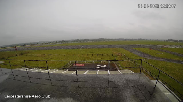 A view from the Leicestershire Aero Club webcam, showing a large grass airfield under a cloudy sky. In the foreground, there is a light gray fence surrounding the airfield, with a view of a small red square marking on the ground, and a white X shape made of rocks. The airfield is surrounded by green fields, with a distant line of hills or trees. The sky is overcast, suggesting a dull day with no visible sunlight.