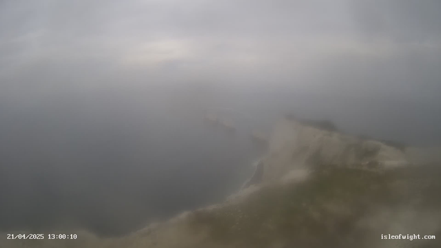A foggy coastal scene with blurred visibility over the sea. The sky is overcast and gray, blending into the mist that envelops the shoreline. The water appears dark and indistinct, with rocky cliffs partially visible in the foreground. The overall atmosphere is calm but obscured by low visibility conditions.