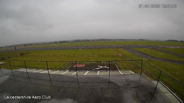 A view from a webcam at Leicestershire Aero Club shows a wide expanse of grassy airfield beneath a cloudy sky. The foreground features a paved area with a cross marking the spot, surrounded by a low fence. In the distance, there are runways and more grasslands, with a few signs visible along the edges. The overall lighting is dim due to overcast weather, and the scene conveys a tranquil, open-air atmosphere.