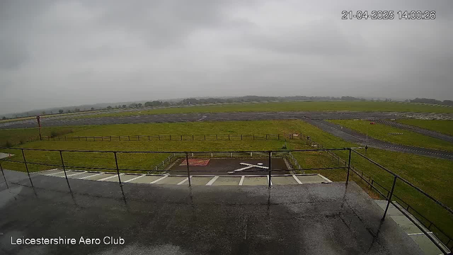 A cloudy sky with gray overcast covers the area. In the foreground, a flat surface is visible with a railing. Below, there is a grassy field that stretches into the distance. An airstrip runs parallel to the foreground, bordered by a wooden fence. Some markings can be seen on the airstrip, along with a red sign. The overall atmosphere appears misty and quiet, indicating overcast weather with limited visibility.