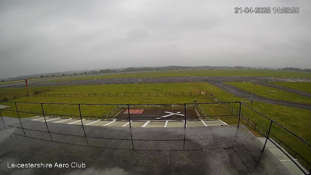 A view from a webcam located at the Leicestershire Aero Club, showing a wide expanse of grass and runways under a cloudy sky. The ground is mostly green with a few patches of dirt. In the foreground, there is a railing surrounding a flat surface, and a large helipad marked with a white "X" is visible. On the left side, a red sign is shown, and two runways extend in the distance, leading to a blurred horizon where trees can be faintly seen. The overall atmosphere is overcast, indicating a potentially rainy day. The time and date are displayed in the top right corner.