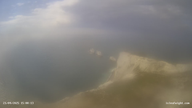 A misty, cloudy view of a coastline with cliffs leading down to the ocean. The water appears calm and reflects the overcast sky. There are distant rock formations off the coast, partially obscured by haze. The date and time are displayed at the bottom left corner.