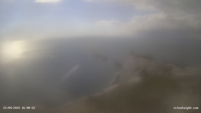 A foggy landscape showing the sea and a distant coastline. The sky is overcast with soft gray clouds. The water has a subtle shimmer reflecting light, while the horizon blends into the fog, obscuring details. The scene conveys a tranquil yet mysterious atmosphere.