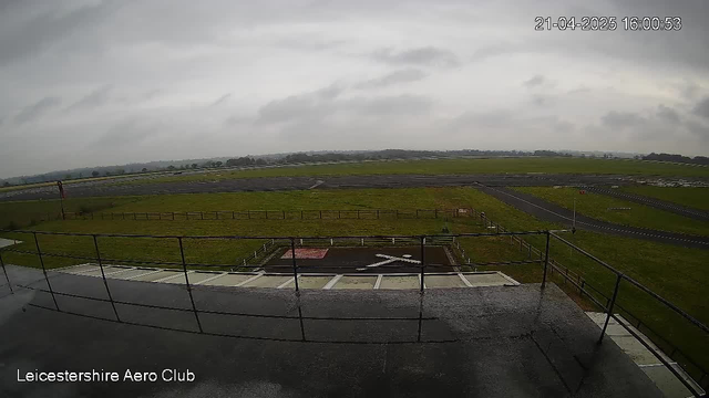 A wide view from a webcam on a cloudy day, showing an airfield. In the foreground, there is a raised platform with a railing. Below, the ground is wet and features a large 'X' marking, likely indicating a landing spot. The airfield extends into the distance, lined with a wooden fence, and is surrounded by green grass. The sky is overcast, adding a grayish hue to the scene.