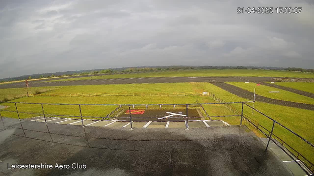 A view from a webcam at Leicestershire Aero Club shows a large open field with a grassy surface and a runway for aircraft. The sky is overcast with gray clouds. In the foreground, there is a railing and a horizontal platform area with white markings resembling a cross. Farther back, there is a fence surrounding part of the field, and in the distance, a few trees can be seen. The runway is visible to the right, with black asphalt and marked pathways.