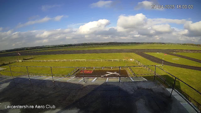 A wide view of a grassy airfield under a partly cloudy sky. In the foreground, a fenced area contains a landing pad marked with a white X. The runway extends into the distance, bordered by green fields and trees. There is a flagpole on the left, with the flag fluttering in the breeze. The scene is bright and well-lit, indicating daytime.
