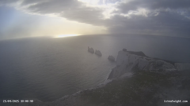 A coastal scene at sunset, depicting the ocean stretching to the horizon under a cloudy sky. In the foreground, there are rocky cliffs and outcrops of land, with three prominent rock formations jutting out into the water. One of the formations features a small building or structure. The sunlight is reflecting off the ocean, creating a shimmering effect on the water's surface. The overall mood is tranquil and serene.