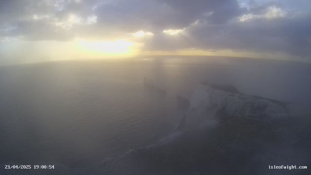 A coastal landscape featuring cliffs and the ocean under a cloudy sky. The sun is setting on the horizon, casting a warm, golden light over the scene. Mist or haze is present, softening the view of the cliffs and the water. The foreground shows rocky cliffs meeting the sea, with gentle waves lapping against them. The date and time are displayed in the lower left corner.