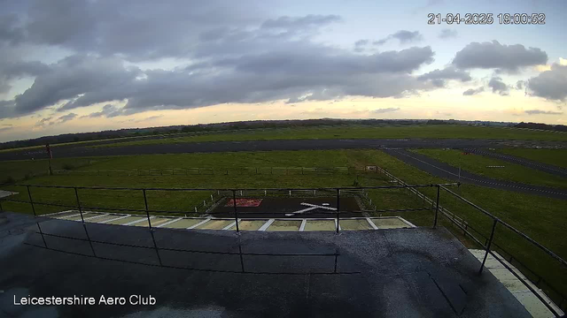 A view from a rooftop at Leicestershire Aero Club, showcasing a wide expanse of an airfield. The foreground features a railing with a wet surface, and beyond it, a grassy area with a marked section on the ground resembling a cross. In the midground, there is a paved runway stretching across the field, bordered by green grass and a few scattered trees in the background. The sky is partly cloudy, with soft shades of orange and blue indicating sunset. The time displayed is 19:00 on April 21, 2025.