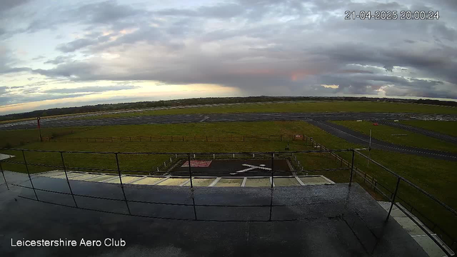 A panoramic view of an airfield at dusk, featuring a grassy runway and taxiways. In the foreground, a safety railing lines the observation deck of a building labeled "Leicestershire Aero Club." The sky is partly cloudy with soft hues of blue, pink, and orange as the sun sets. The landscape is mostly flat with distant trees visible at the horizon.