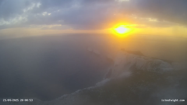 A hazy view of the ocean during sunset, with the sun casting a bright orange glow on the horizon. The sky is filled with gray and soft pastel colors, while the ocean appears dark and indistinct. There is a faint outline of cliffs on the right side of the image, partially shrouded in mist. The date and time are displayed in the lower left corner.