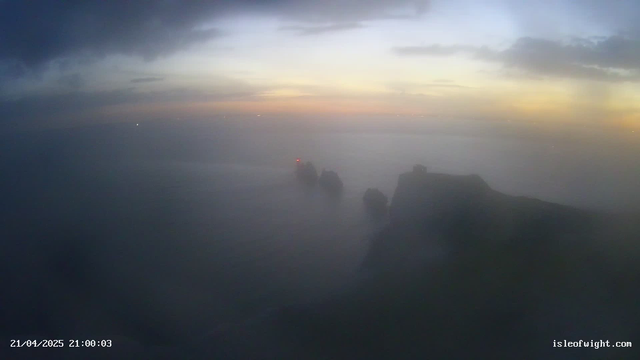 A foggy view of the coastline at dusk, featuring dark shapes of rocks jutting out from the water. A lighthouse with a red light is visible among the rocks, while the sky transitions from dark to lighter hues near the horizon, suggesting sunset. The overall atmosphere is misty and unclear, with soft ambient colors blending in the background.