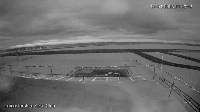 A black and white image showing a view from a webcam overlooking the Leicestershire Aero Club. The foreground features a flat rooftop area with a fence and markings that suggest an airstrip or helipad. In the background, a wide view of the landscape is visible, with dark clouds in the sky and a body of water extending into the distance. The image reflects a somber, cloudy atmosphere with minimal light.