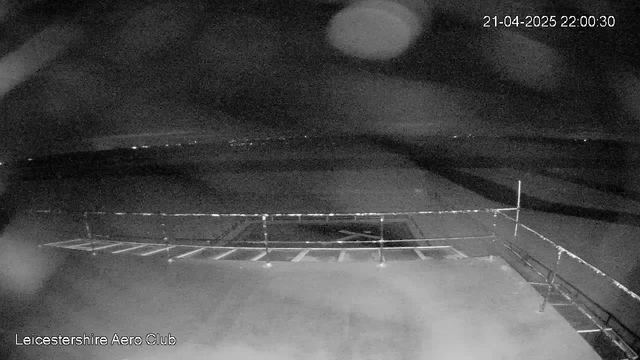 A dark webcam image showing a view from a high platform or deck. In the foreground, there is a railing that borders the deck, with several horizontal bars visible. The background features a dark landscape with faint lights in the distance, suggesting a distant horizon or city lights. The overall scene is dimly lit, indicating that it is nighttime.