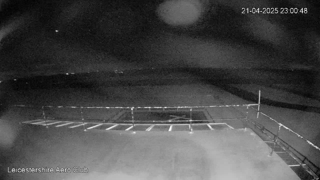A dimly lit scene from a webcam showing an outdoor area at night. The foreground features a railing with a series of horizontal bars, while the background displays a flat landscape with a cloudy sky. Small lights are visible in the distance, suggesting additional structures or activity, but overall the image is predominantly dark. A timestamp in the upper right corner indicates the date and time as April 21, 2025, at 11:00 PM. The bottom left corner identifies the location as Leicestershire Aero Club.