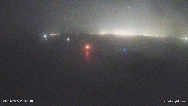 A foggy scene over water at night, with a few distant lights visible along the horizon. One prominent red light and several smaller blue lights reflect on the water's surface. The overall image is dimly lit with limited visibility due to fog. The timestamp at the bottom indicates the date and time as April 21, 2025, at 11:00 PM.