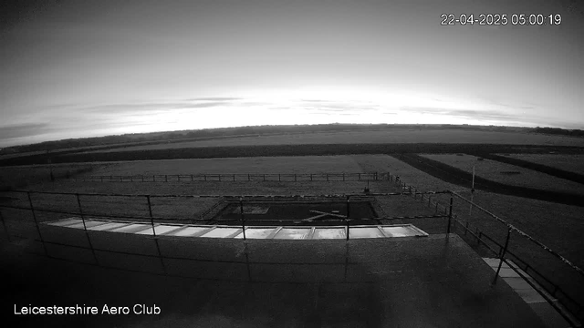 A black and white image of a landscape viewed from a high vantage point. In the foreground, there is a flat rooftop terrace with glass panels. Below, a runway marked with an "X" is visible, surrounded by grass and a fenced area. The background features a horizon with a cloudy sky as the sun rises, casting a soft light over the scene. The image captures the early morning stillness at the Leicestershire Aero Club.