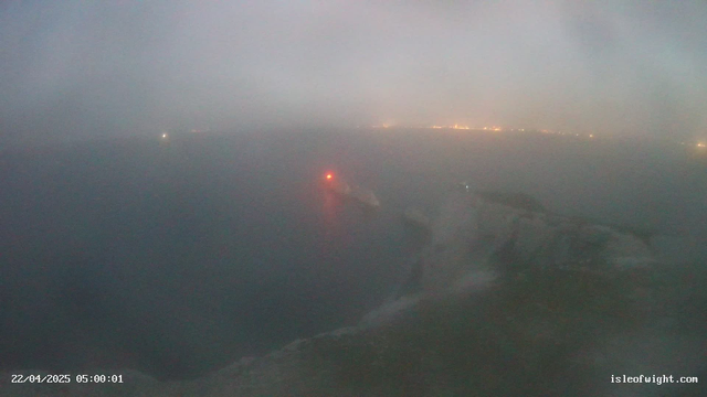 A foggy scene over a body of water with a faint view of a cliff or shoreline on the right. A small red light is visible on a distant object, possibly a buoy or a boat. The atmosphere appears hazy, limiting visibility. In the background, there are dim lights suggesting a distant coastal town or city. The time and date in the corner indicate early morning on April 22, 2025.