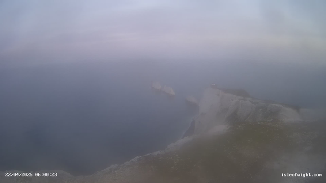 A foggy coastal scene features a rocky cliff on the right side, with the ocean visible below. The colors are muted, with gray and blue tones dominating. In the background, several indistinct shapes resembling boats are partially obscured by the mist. The overall atmosphere is hazy and serene, suggesting an early morning setting.