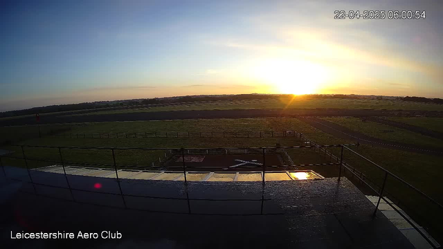 A view of a landscape at sunrise, with the sun rising on the horizon illuminating the sky with warm orange and yellow hues. In the foreground, a railing is visible, with a portion of the ground and a tarmac area partially covered by grass. A marked area on the ground is visible, likely a helipad or runway, with a cross shape formed by white lines. The distance shows an expansive grassy area and possibly more tarmac, under a clear sky. The timestamp at the top indicates the date and time.