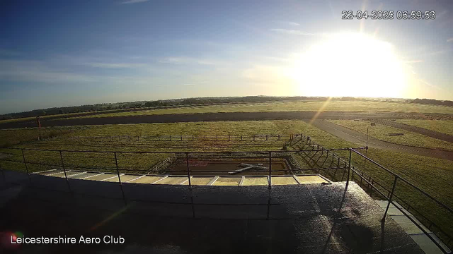 A panoramic view from a high vantage point at Leicestershire Aero Club. The scene features a sun rising over lush green fields and a clear blue sky. A runway is visible in the distance, with a few scattered trees along the horizon. In the foreground, there is a railing and a concrete pathway leading to the edge of the viewing area, and a grassed area is visible below. The image captures a tranquil and expansive landscape during early morning.