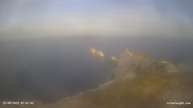 A hazy view of a cliffside overlooking the ocean. The scene features rocky outcrops extending into the water, with a gentle gradient of colors in the sky ranging from light blue to pale gray. The foreground shows a grassy area with scattered flowers, while the background features the shimmering surface of the water blending into the misty horizon. The date and time are displayed in the corner.
