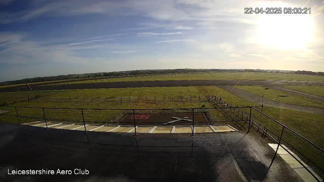 A view from a webcam shows a grassy area with a small runway. In the foreground, there is a wooden railing and a terrace that leads to an area marked with an "X" on the ground. The scene is bathed in morning light with a bright blue sky and some wispy clouds. On the horizon, fields stretch out, indicating a rural landscape. The time stamp at the top right shows it was taken at 08:00:21 on April 22, 2025.