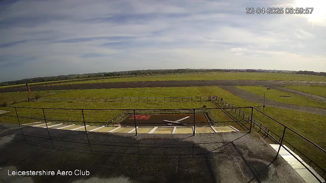 A view from a webcam at Leicestershire Aero Club, capturing a grassy runway area with a large expanse of green grass and a blue sky scattered with white clouds. In the foreground, a railing runs along the edge of a building. There is a small ramp leading to the runway, which appears empty. In the distance, the runway and landscape continue, with a few trees and structures visible on the horizon. The time and date displayed at the top right reads 22-04-2025 08:59:57.