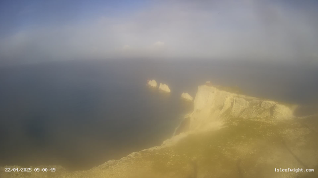 A coastal scene viewed from a high vantage point. In the foreground, there are rugged cliffs that slope down towards the water. The cliffs have a pale color and are partially bathed in sunlight. Below, the ocean appears calm and has a gradient of deep blue to lighter shades near the shore. Three large rock formations protrude from the water, with some clouds visible in the distance. The atmosphere is slightly hazy, giving the scene a soft appearance. In the top right corner, a small building is situated on the cliff's edge, near a pathway leading away from it. The image is time-stamped, showing a date and time.