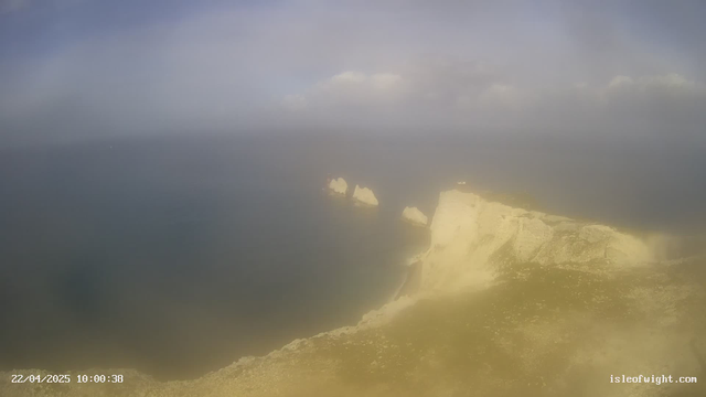 A coastal scene features a rocky shoreline with steep, white cliffs rising from the sea. The water is calm and extends towards the horizon, blending into a hazy blue sky filled with soft, white clouds. A few rock formations jut out from the water near the center of the image. The lower part of the image shows grassy terrain atop the cliffs, with some scattered stones visible.