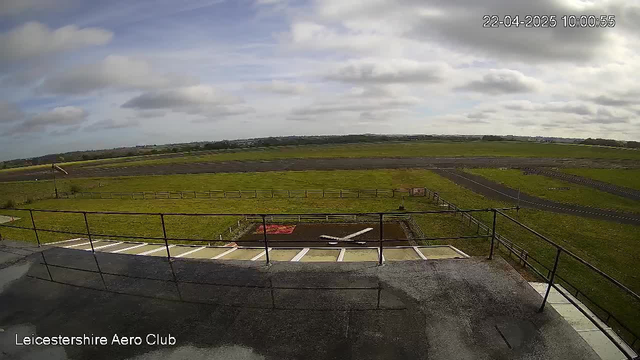 The image shows a view from a webcam at Leicestershire Aero Club. In the foreground, there is a flat rooftop with a railing. Below the roof, a small model airplane is seen on the ground. The background features a large grassy field and a runway, with a few scattered clouds in a mostly blue sky. Some trees and distant hills are visible in the horizon, and the overall atmosphere is serene and open. The time displayed in the corner reads 10:00 AM on April 22, 2025.